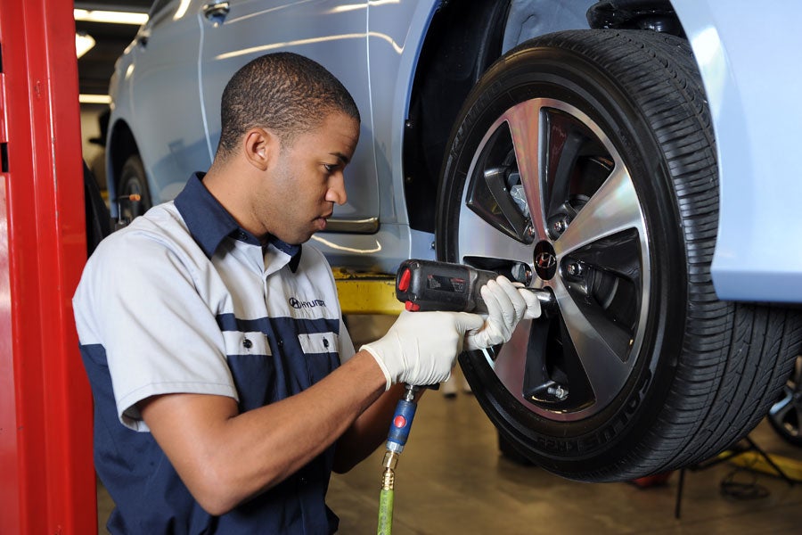 A mechanic wearing a two-toned uniform and gloves removes a car tire with an air impact wrench.