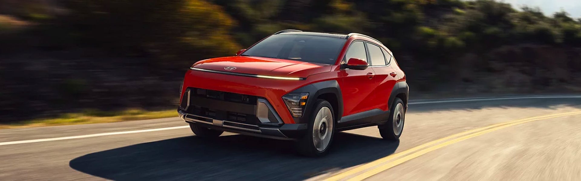 A red Hyundai Kona EV drives on a winding road with blurred green foliage in the background.