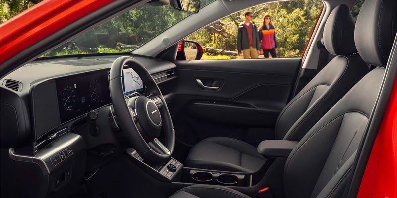 View of a modern car's black interior, showing the dashboard, steering wheel, and front seats. Two hikers are visible through the open passenger door.
