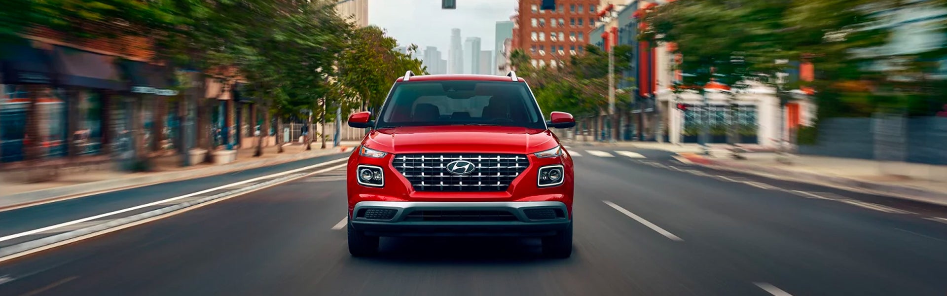 A Hyundai Venue car drives down a city street with blurred buildings and trees on either side.