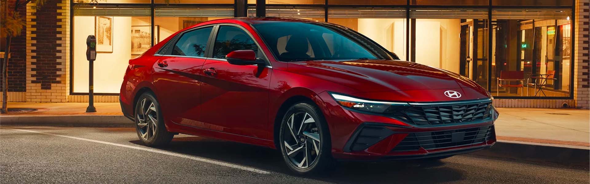 Hyundai Elantra parked on a city street at night, with a brick building and brightly lit storefront in the background.