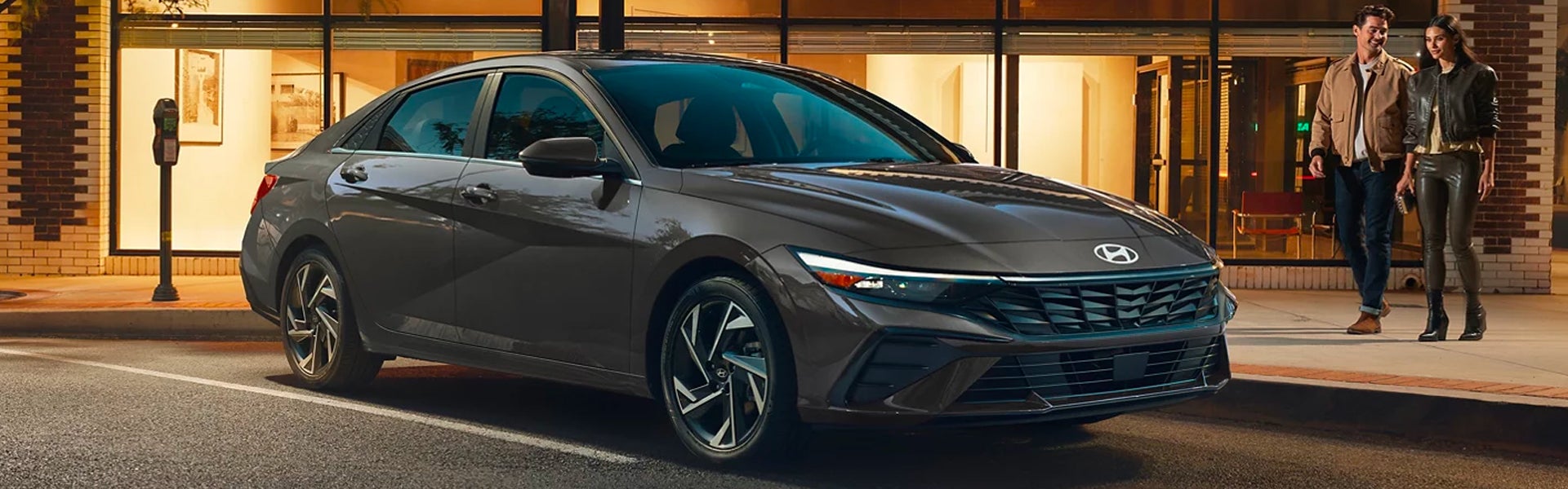 A brown Hyundai Elantra is parked by a sidewalk with a couple walking in the background.