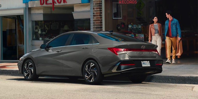 A dark gray sedan parked on a city street, with two people walking in the background.