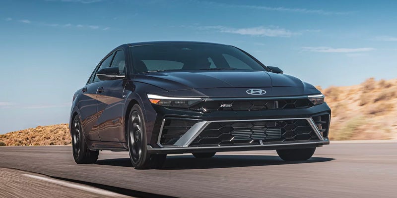 Front view of a dark gray Hyundai Elantra N Line car in motion on a road, with desert hills in the background.