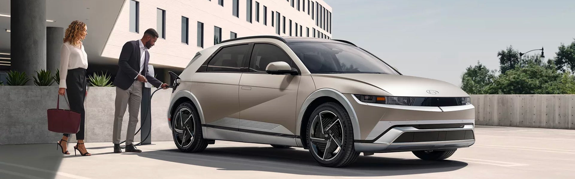 A man plugs a charger into a tan electric car while a woman stands nearby holding a large purse.