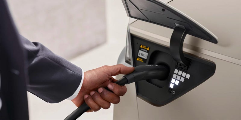 A hand in a dark suit plugs a charging cable into an electric vehicle.