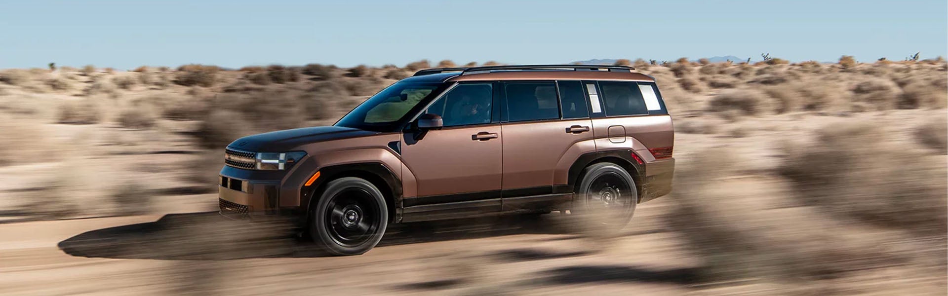SUV drives fast across a dirt road, kicking up dust. The background is a blurry desert landscape.