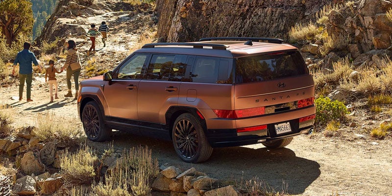A family of four walks up a rocky dirt path next to a bronze Hyundai Santa Fe SUV.