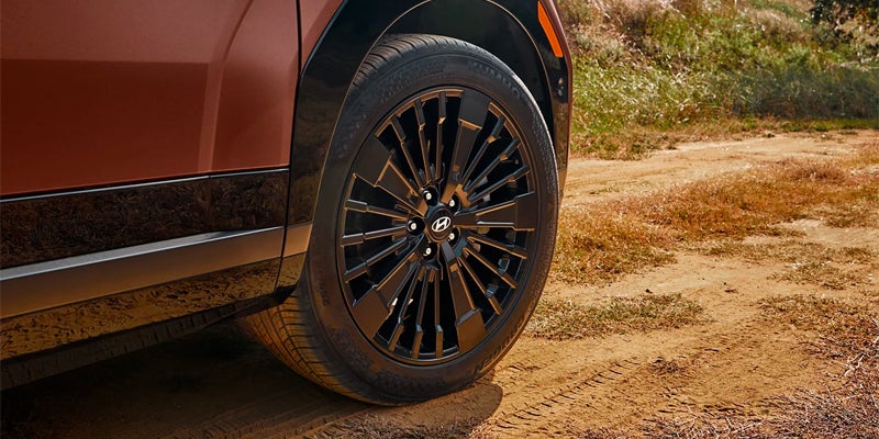 Close-up of a car's front wheel with a multi-spoke, glossy black rim on a dirt road.
