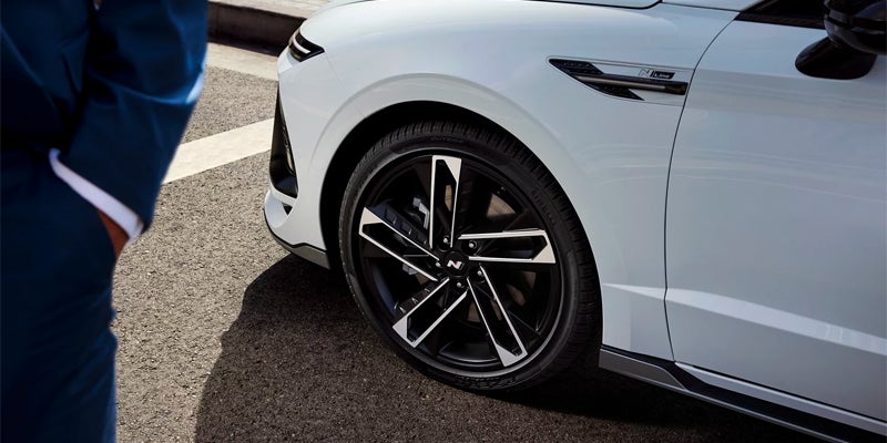 A person in a blue suit stands next to the front wheel of a Hyundai car. The wheel has black and silver rims.