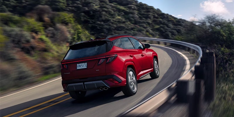 Rear view of a Hyundai Tucson SUV driving around a bend on a road with green hills.