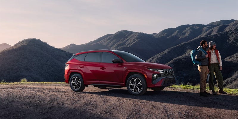 A Hyundai Tucson SUV parked on a dirt road, facing mountains. Two hikers stand by the car, looking at the view.