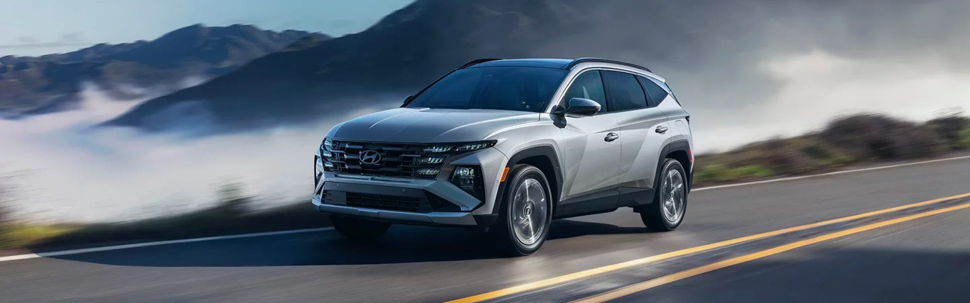 Hyundai Tucson SUV drives on a road with mountains and clouds in the blurred background.