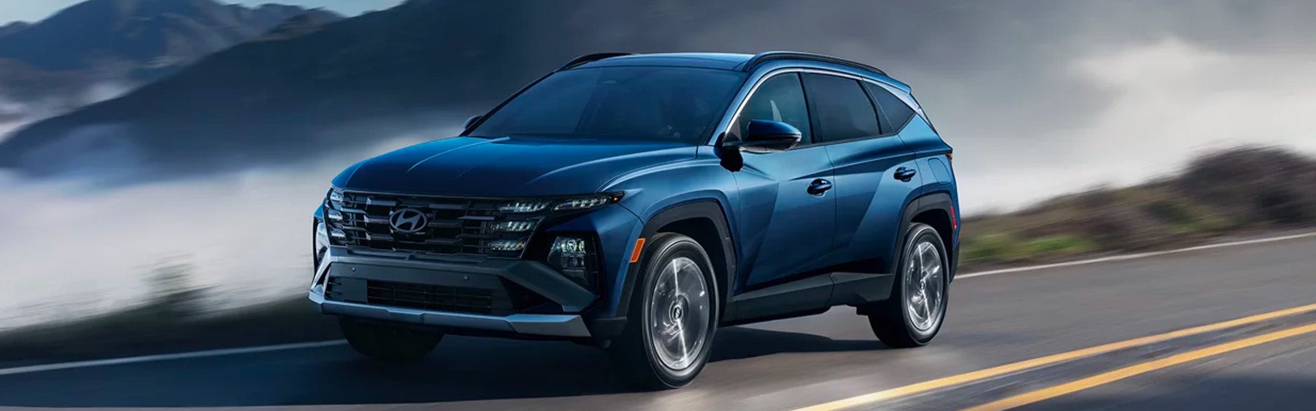 A Hyundai Tucson drives on a road with mountains and fog in the background.