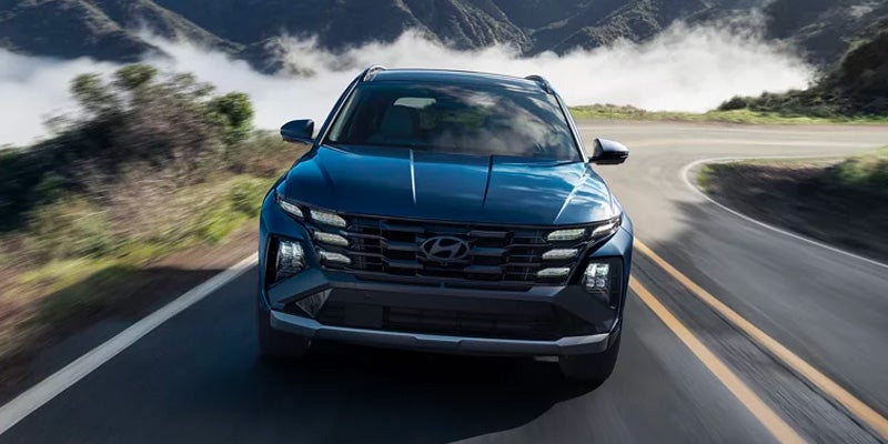 A Hyundai Tucson SUV drives on a winding road with mountains and clouds in the background.