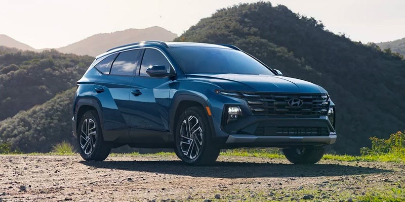 Front three-quarter view of a Hyundai Tucson SUV parked on a dirt road with green mountains in the background.
